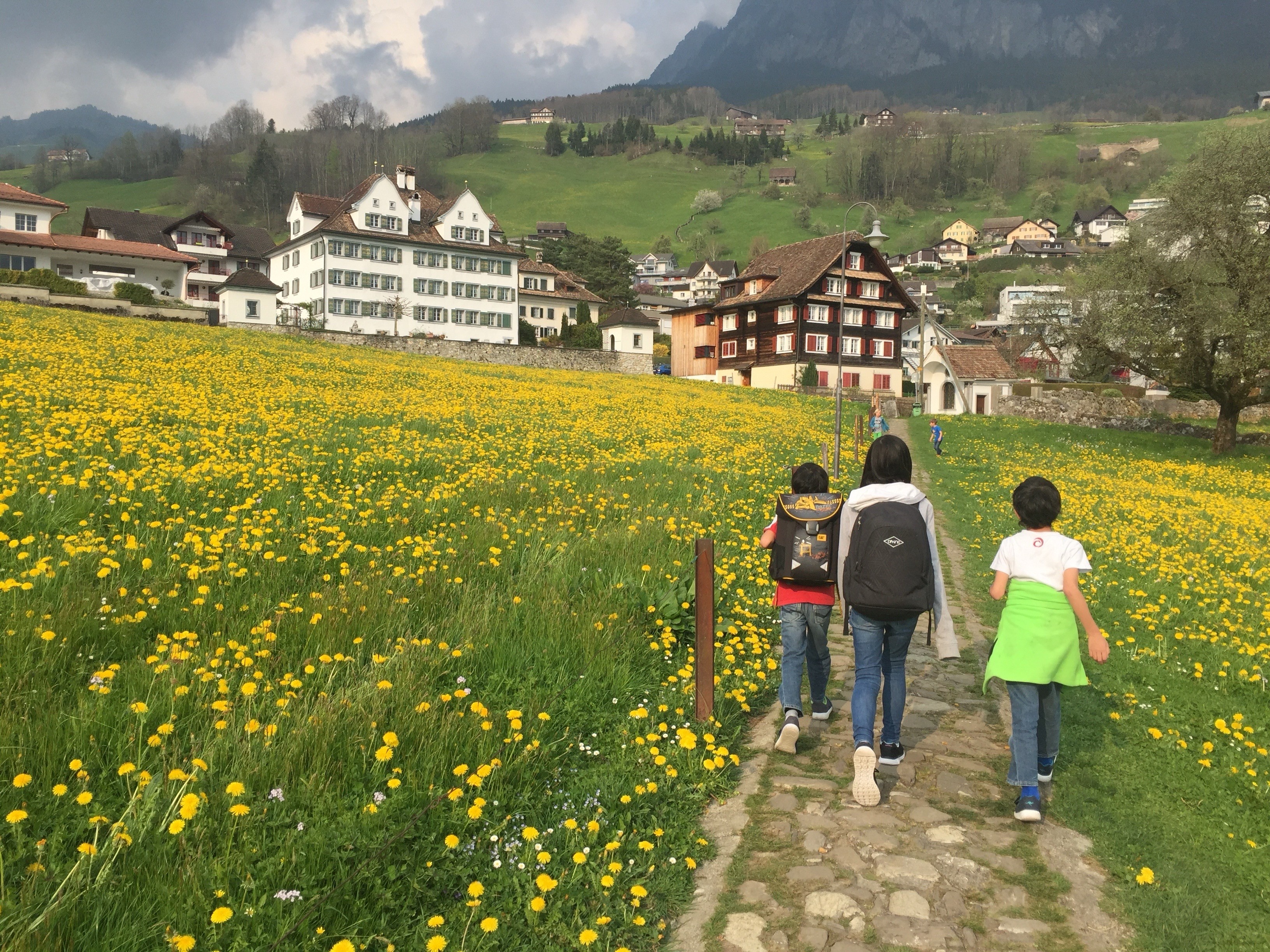 Drei Kinder gehen über einen sehr unebenenalten Steinweg durch eine Wiese mit vielen glben Blumen. Im Hintergrund sind einige Häuser zu sehen, unter anderem ein grosses, älteres Herrenhaus. Drei Kinder gehen über einen sehr unebenenalten Steinweg durch eine Wiese mit vielen glben Blumen. Im Hintergrund sind einige Häuser zu sehen, unter anderem ein grosses, älteres Herrenhaus.