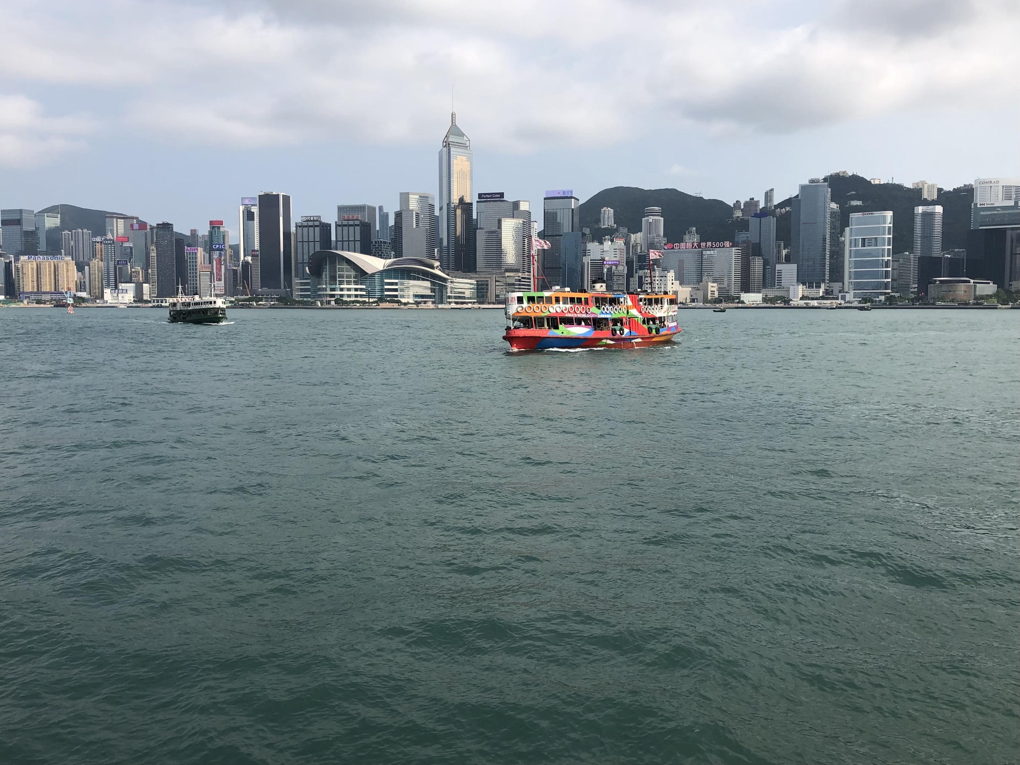 Zwei Hafenfähren, eine davon bunt bemalt vor der Skyline von Hong Kong Island. Im Hintergrund sind die bewaldeten Hügel der Insel zu sehen. Zwei Hafenfähren, eine davon bunt bemalt vor der Skyline von Hong Kong Island. Im Hintergrund sind die bewaldeten Hügel der Insel zu sehen.