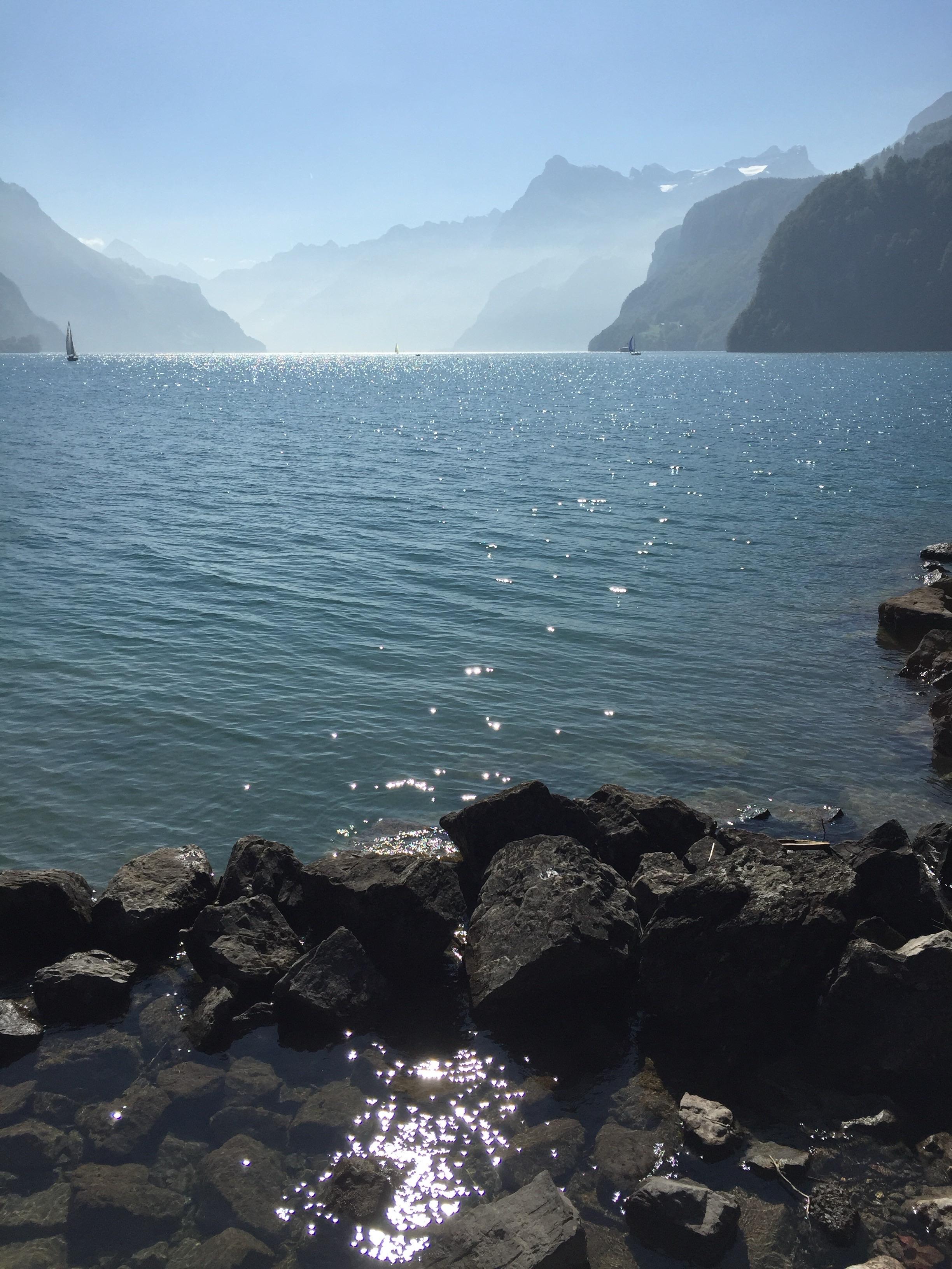 Vor im schwachen Dunst stehenden Bergen und blauem Himmel ist ein ziemlich ruhiger See zu sehen. Weit weg segeln zwei Segelboote. Im Vordergrund liegen dunkle Steine eines Wellenbrechers und im Wasser spiegelt sich dutzendfach die Sonne als kleine glänzende Punkte. Vor im schwachen Dunst stehenden Bergen und blauem Himmel ist ein ziemlich ruhiger See zu sehen. Weit weg segeln zwei Segelboote. Im Vordergrund liegen dunkle Steine eines Wellenbrechers und im Wasser spiegelt sich dutzendfach die Sonne als kleine glänzende Punkte.